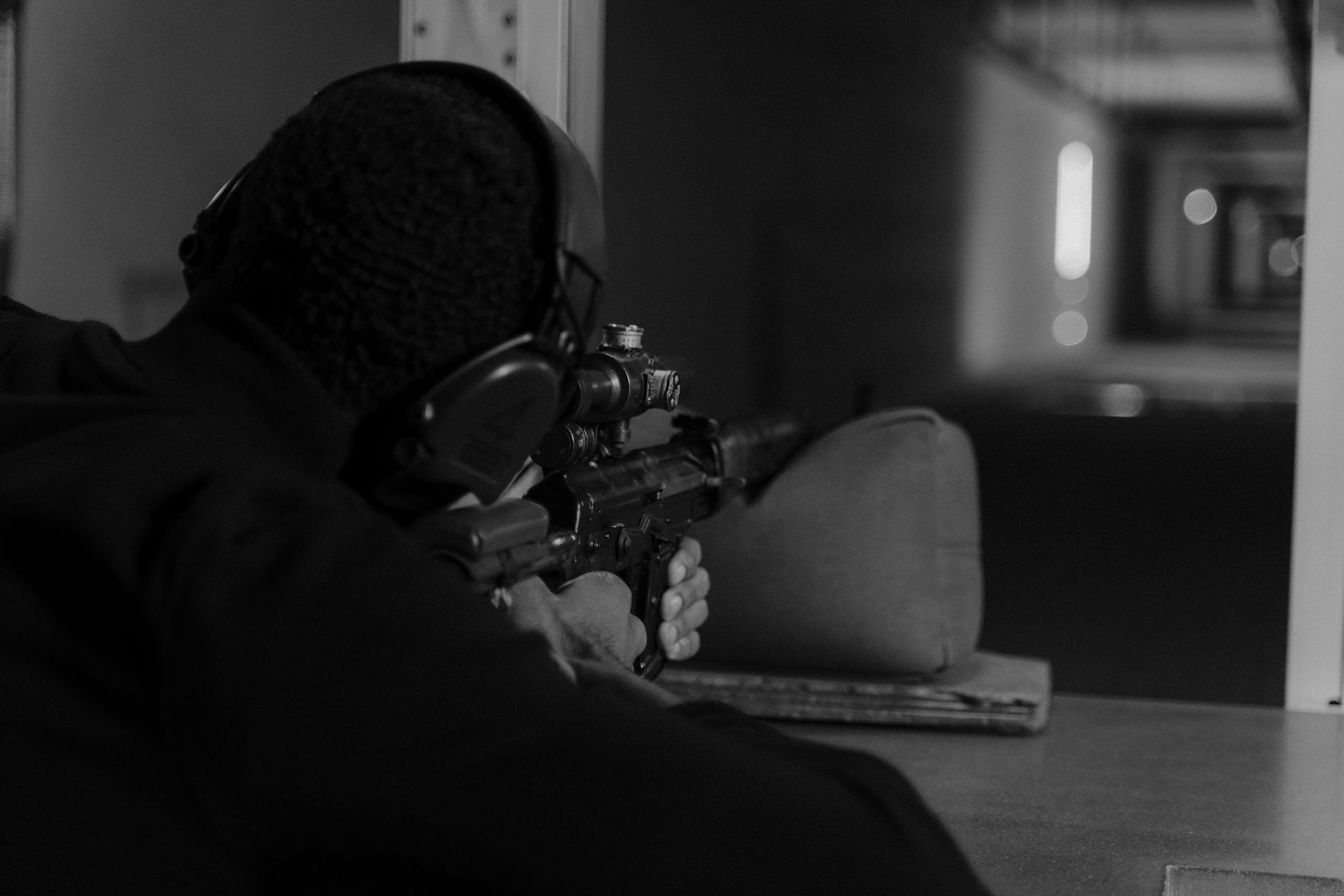Black and white photo of a man aiming a rifle at an indoor shooting range.