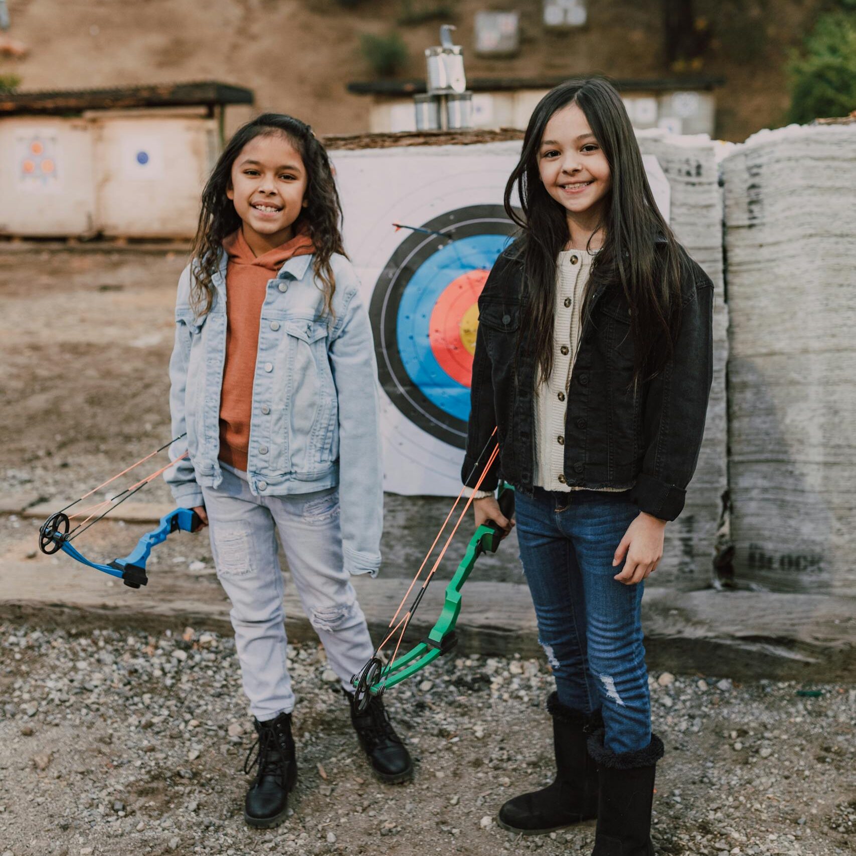 Two young girls with bows stand near an archery target outdoors.
